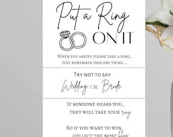 以下が含まれることがあります： 「Put a Ring On It」というテキストとゲームの指示が書かれた白黒の印刷可能なサイン。サインには、ダイヤモンドが付いた2つの絡み合ったリングのシンプルなイラストが描かれています。