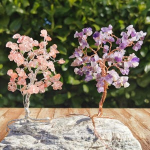 Peut inclure: Deux arbres décoratifs en pierres précieuses. L'un présente des pierres roses avec un tronc et des branches en fil d'argent. L'autre a des pierres violettes avec un tronc et des branches en fil de cuivre. Les deux arbres sont montés sur une base en pierre grise.