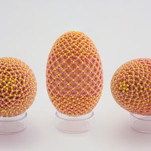以下が含まれることがあります： ピンクと黄色のビーズでできた装飾的なイースターエッグが3つ。卵は透明なスタンドに飾られています。