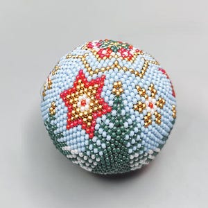 May include: A round, beaded ornament with a light blue background. The ornament features a red star, gold accents, and green and white floral designs. The beadwork is intricate and detailed, creating a textured surface.