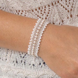 Puede incluir: Una pulsera blanca con detalles de perlas. La pulsera se lleva en la muñeca y presenta varias filas de pequeñas cuentas blancas y perlas redondas más grandes. La pulsera se coloca sobre un fondo de un suéter de punto abierto color crema.