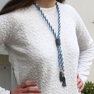 May include: A blue and white twisted rope necklace with a dark square clasp and tassel pendant. The necklace is worn over a white quilted long-sleeved top. The background is a white wall and a small green bush.
