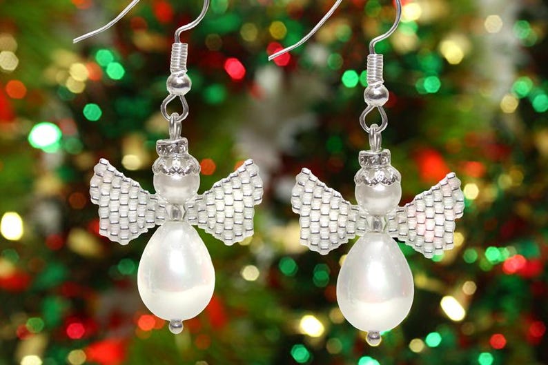 May include: A pair of silver angel-shaped earrings with white pearl accents. The earrings are hanging from a Christmas tree.