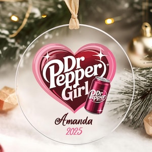 May include: A clear, round Christmas ornament with a Dr. Pepper Girl design inside a pink heart. The ornament includes a Dr. Pepper can graphic, the name "Amanda" and the year "2025". The ornament hangs from a beige ribbon.