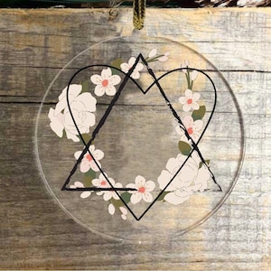 May include: Clear, round Christmas ornament with a black heart and star design, filled with white flowers and green leaves. The ornament hangs from a gold and black ribbon. Festive holiday decor.