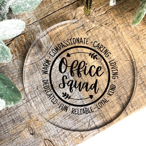 May include: A clear, round ornament with the words "Office Squad" in a black, circular design. The ornament is surrounded by words like "compassionate" and "loving." It hangs from a gold string, set against a rustic wooden background with green foliage.