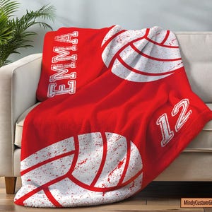 May include: A red fleece blanket featuring a white distressed volleyball graphic, the name EMMA printed vertically in white block letters, and the number 12 in white. The design has a textured, worn appearance across the fabric surface.