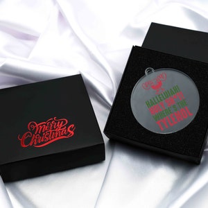 May include: A black gift box with the words "Merry Christmas" in red script. Next to it, an open black box reveals a clear, round Christmas ornament with the text "Hallelujah! Holy S***! Where's the Tylenol?" in red and green.