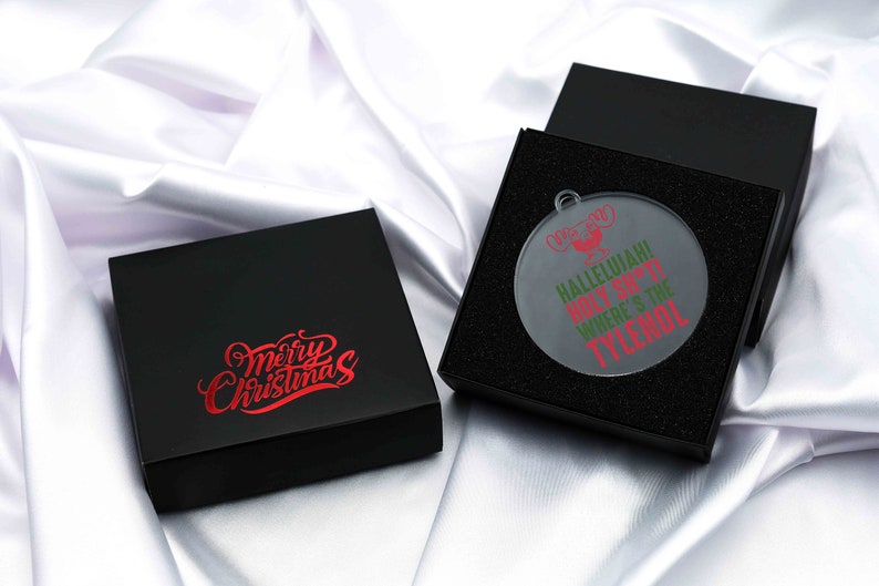 May include: A black gift box with the words "Merry Christmas" in red script. Next to it, an open black box reveals a clear, round Christmas ornament with the text "Hallelujah! Holy S***! Where's the Tylenol?" in red and green.