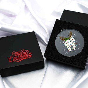 May include: A clear, round Christmas ornament featuring a cartoon tooth decorated with reindeer antlers and Christmas lights. The ornament is displayed in a black gift box, with a second box nearby. The text "Merry Christmas" is printed on the box.