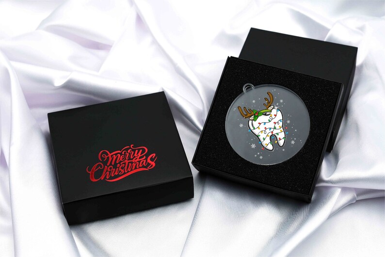 May include: A clear, round Christmas ornament featuring a cartoon tooth decorated with reindeer antlers and Christmas lights. The ornament is displayed in a black gift box, with a second box nearby. The text "Merry Christmas" is printed on the box.