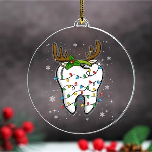 May include: A clear, round Christmas ornament featuring a cartoon tooth decorated with reindeer antlers, holly, and colorful Christmas lights. The ornament has a gold string for hanging and is decorated with snowflakes.