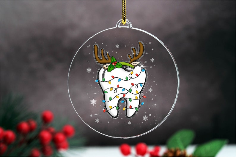 May include: A clear, round Christmas ornament featuring a cartoon tooth decorated with reindeer antlers, holly, and colorful Christmas lights. The ornament has a gold string for hanging and is decorated with snowflakes.