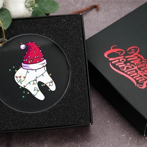 May include: A clear, round Christmas ornament featuring a cartoon tooth wearing a Santa hat and festive lights. The ornament is displayed in a black gift box. The box next to it has the words "Merry Christmas" in red script.