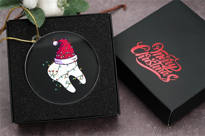 May include: A clear, round Christmas ornament featuring a cartoon tooth wearing a Santa hat and festive lights. The ornament is displayed in a black gift box. The box next to it has the words "Merry Christmas" in red script.