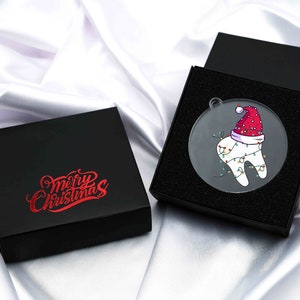 May include: A clear, round Christmas ornament featuring a cartoon tooth wearing a Santa hat and string lights, presented in a black gift box. The box lid displays "Merry Christmas" in red script. The ornament is packaged on a white satin surface.