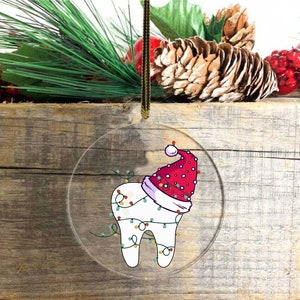 May include: A clear, round Christmas ornament featuring a cartoon tooth wearing a Santa hat and wrapped in Christmas lights. The ornament hangs from a gold-colored string against a rustic wooden background, with pine and berries.