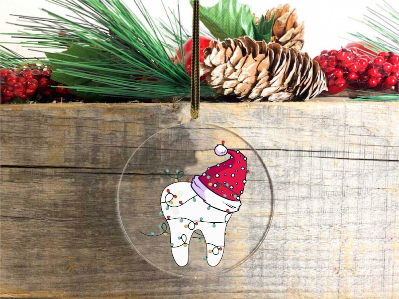 May include: A clear, round Christmas ornament featuring a cartoon tooth wearing a Santa hat and wrapped in Christmas lights. The ornament hangs from a gold-colored string against a rustic wooden background, with pine and berries.