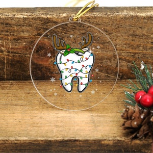 May include: A clear, round Christmas ornament featuring a cartoon tooth decorated as a reindeer. The tooth has brown antlers, holly, and is wrapped in colorful Christmas lights. A gold string is attached for hanging.
