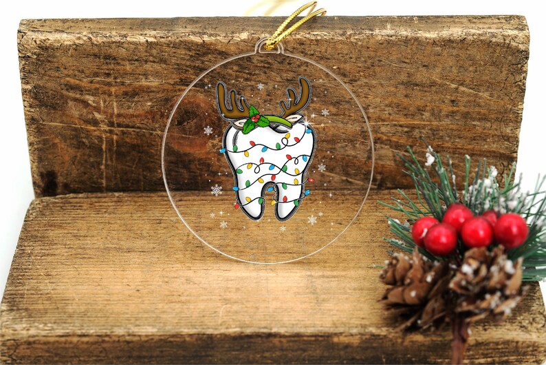 May include: A clear, round Christmas ornament featuring a cartoon tooth decorated as a reindeer. The tooth has brown antlers, holly, and is wrapped in colorful Christmas lights. A gold string is attached for hanging.