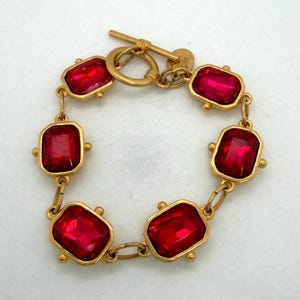 May include: A gold-tone chain bracelet featuring five rectangular, faceted red stones set in metal frames. The bracelet is secured with a toggle clasp and includes a small circular metal tag attached near the closure mechanism.
