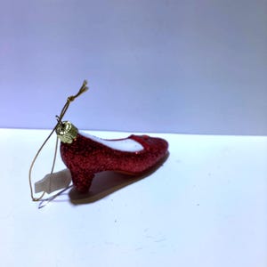 May include: A red glittery high-heeled shoe Christmas ornament with a gold-colored loop for hanging. The shoe has a white interior and a small red bow on the front. The ornament is on a white surface.