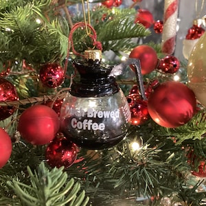 Puede incluir: Adorno navideño de cristal con forma de cafetera, con la inscripción "Fresh Brewed Coffee". El adorno es negro y transparente, colgado entre adornos rojos y dorados y agujas de pino verdes.