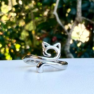 May include: A silver-colored ring with a cat design. The ring features a cat's head with pointed ears and a curled tail. The ring is on a white surface with a blurred green background.