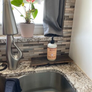 May include: A white bottle of Meyer's Clean Day hand soap with a pump dispenser sits on a wooden soap dish. The soap dish is on a granite countertop next to a stainless steel kitchen sink.