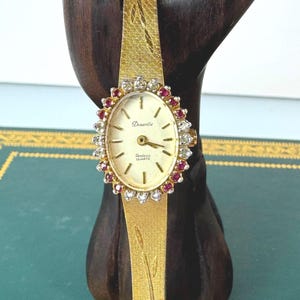 May include: A gold-tone quartz wristwatch with an oval face, featuring a bezel set with alternating clear stones and red stones. The watch has a textured gold-tone metal band with an engraved leaf pattern and a simple cream-colored dial with hands.