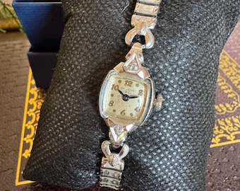 May include: A vintage Bulova watch with a silver band and a rectangular face. The watch face is cream-colored with black numerals and hands. The watch is displayed on a black textured pillow.