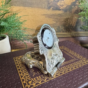 May include: A silver-toned, ornate desk clock with a white clock face and black hands. The clock has a decorative, scrolled design and sits on a hinged base. The clock is displayed on a dark brown, textured surface.