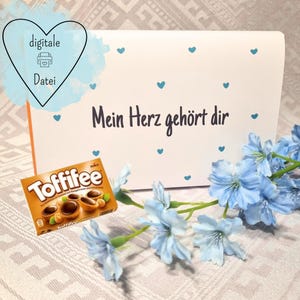 May include: A white gift box with blue heart accents and the German text "Mein Herz gehört dir." A Toffifee candy box and blue flowers are in the foreground. The image also contains the text "digitale Datei."