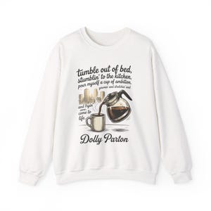 May include: A white crewneck sweatshirt featuring a graphic print of a coffee pot pouring into a mug, a city skyline, and the text: tumble out of bed, stumblin' to the kitchen, pour myself a cup of ambition, yawnin' and stretchin' and and tryin' come to life. Dolly Parton.