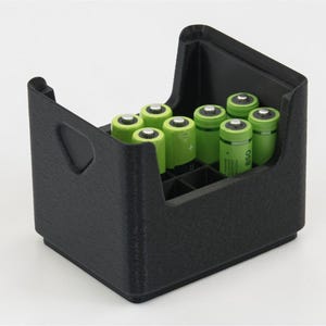 Puede incluir: Un organizador de baterías negro que contiene varias pilas AA verdes. El organizador tiene una superficie texturizada y un recorte en forma de asa en el lateral. Las baterías están ordenadas dentro del contenedor.