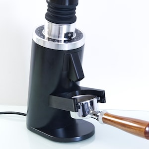 Può includere: Un macinacaffè elettrico nero con quadrante argentato e manico in legno. Il macinacaffè ha un portafiltro e un soffietto in gomma nera. Il design è elegante e moderno, adatto all'uso domestico o commerciale.