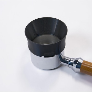 Könnte beinhalten: Ein schwarzer Kaffeefiltertrichter sitzt auf einem Edelstahl-Siebträger mit Holzgriff. Der Trichter soll verhindern, dass Kaffeepulver verschüttet wird. Das Bild ist vor einem weißen Hintergrund aufgenommen.