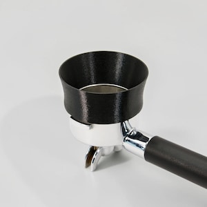 May include: A black espresso portafilter funnel attachment sits atop a chrome-plated metal portafilter basket. The funnel features a wide, flared opening designed to guide coffee grounds into the basket, attached to a black handle.