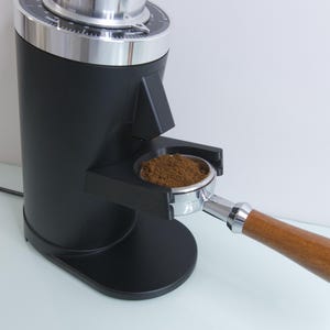 Può includere: Un macinacaffè elettrico nero con la parte superiore argentata e un portafiltro con chicchi di caffè macinati. Il macinacaffè ha un tamper integrato.