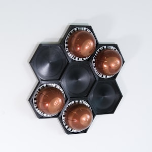 May include: A black, hexagonal coffee pod holder with six compartments. Four compartments hold bronze-coloured coffee pods with white and black striped rims. The holder is mounted on a white surface.