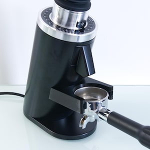 May include: A black electric coffee grinder with a silver top and a portafilter holder. The grinder has a dial with numbers and a black handle. The portafilter is silver with a black handle. The grinder is on a white surface.