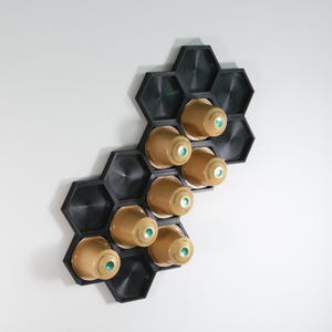 May include: A black, hexagonal coffee pod organiser fixed to a white wall. It holds several gold-coloured coffee pods with green detailing. The design is modern and space-saving, ideal for a kitchen or coffee area.