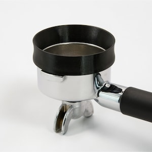 Puede incluir: Un portafiltro de café negro con una cesta de filtro de acero inoxidable. El portafiltro tiene un mango negro y un anillo de goma negro alrededor de la parte superior. Diseñado para máquinas de espresso.