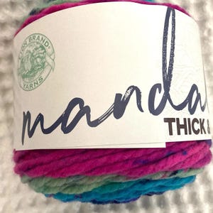 May include: A skein of Lion Brand Mandala Thick & Quick yarn in a multi-colored pattern with pink, blue, and green hues. The label reads "mandala" and "Thick & Quick".