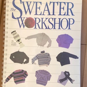 May include: A spiral-bound book titled "The Sweater Workshop" by Jacqueline Fee. The cover features a large blue title with a yarn ball and illustrations of various sweaters in different colors and styles, including a striped sweater and a turtleneck.