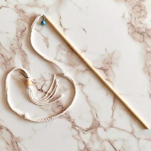 May include: A cat toy featuring a natural wooden stick with a braided white rope and a small blue bell. The rope has a wooden bead and tassels. The toy is designed for interactive play with cats.