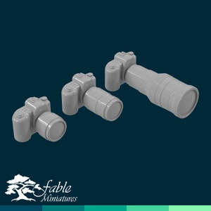 Puede incluir: Tres cámaras de juguete de plástico gris de diferentes tamaños. Las cámaras están dispuestas en una fila de la más grande a la más pequeña. La cámara más grande tiene un objetivo largo. Las cámaras están fabricadas por Fable Miniatures.