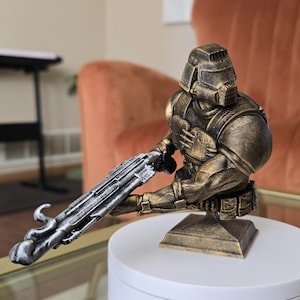 May include: A bronze-colored bust of a futuristic soldier figure, holding a silver and black weapon. The figure is mounted on a square base and displayed on a white, round platform. The bust is a decorative item.