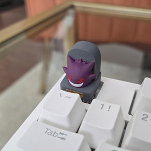 May include: A custom keycap featuring a purple Gengar character on a gray tombstone design. The keycap is placed on a white mechanical keyboard, showcasing a unique accessory for gamers and keyboard enthusiasts.