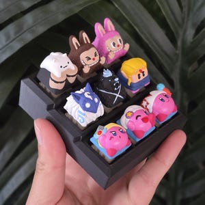 May include: A black keycap holder containing nine colourful, cartoon-style keycaps. The keycaps depict various characters in shades of pink, blue, brown, and white. The holder is held in a hand against a backdrop of green leaves.